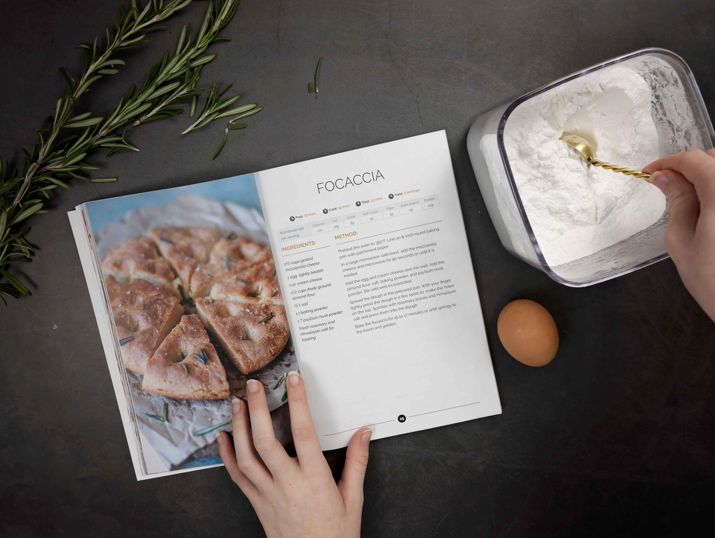 A woman's hand opens the keto carbs cookbook on a grey surface. A bowl of flour in the mixed, an egg and a sprig of rosemary can be seen.