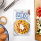 The Paleo Breakfast Recipes Cookbook placed on a plain white surface, accompanied by a bowl of eggs, sea salt, and a cutting board with diced vegetables.