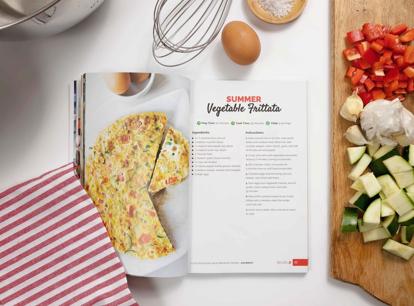 An opened Paleo Breakfast Cookbook placed on a white surface, surrounded by a wooden board with diced vegetables, an egg, salt, a whisk, and a kitchen cloth.