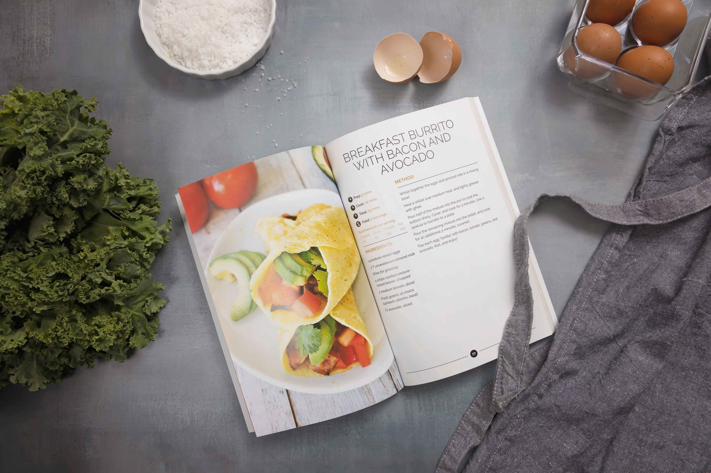 An open Keto breakfast cookbook is placed on a marble surface, alongside eggs, vegetables, and sea salt.