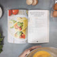 An open Keto breakfast cookbook is placed on a marble surface, alongside eggs, vegetables, sea salt and a cooking pan..