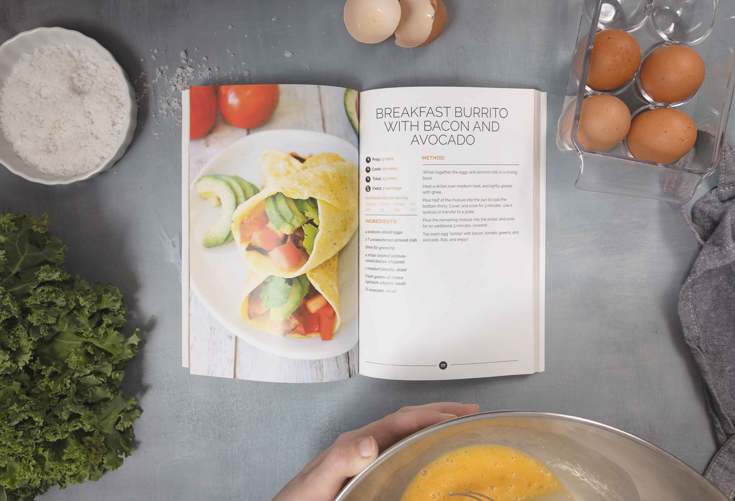 An open Keto breakfast cookbook is placed on a marble surface, alongside eggs, vegetables, sea salt and a cooking pan..