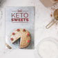 The Keto Sweets Cookbook  placed on a white marble surface. A bowl of flour, cacao powder, and eggs can be seen on the right.