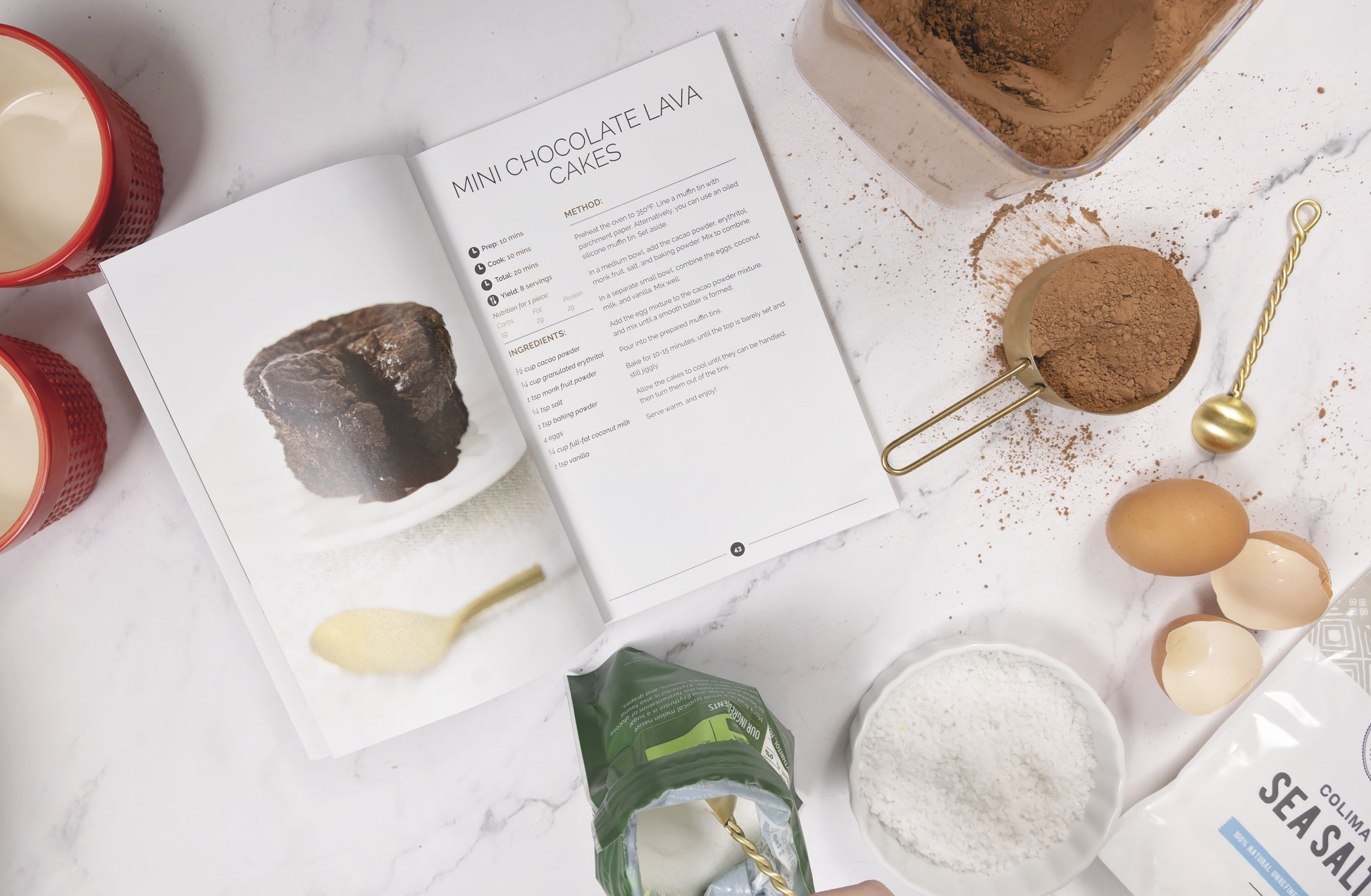 An opened Keto Sweets Cookbook laid on a plain marble surface surrounded by sea salt, chocolate, and egg.