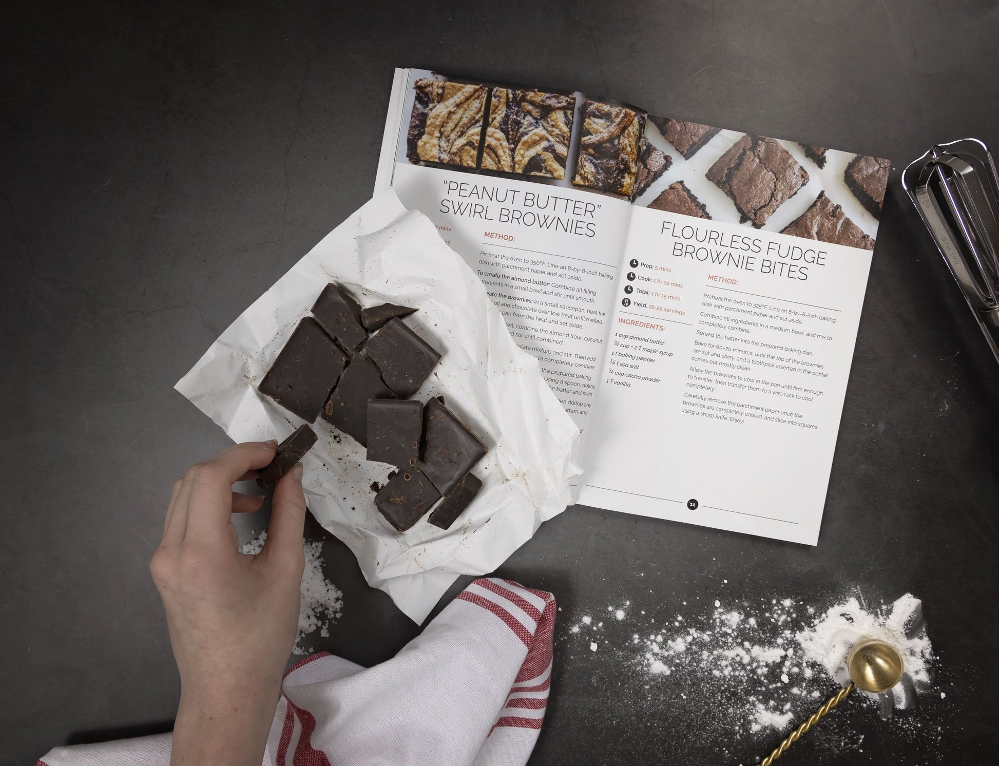A woman's hand picks up the chocolate bars placed on the left-side pages of an open Paleo Sweets Cookbook, which is laid on a plain grey surface. A measuring spoon and a whisk are also visible.