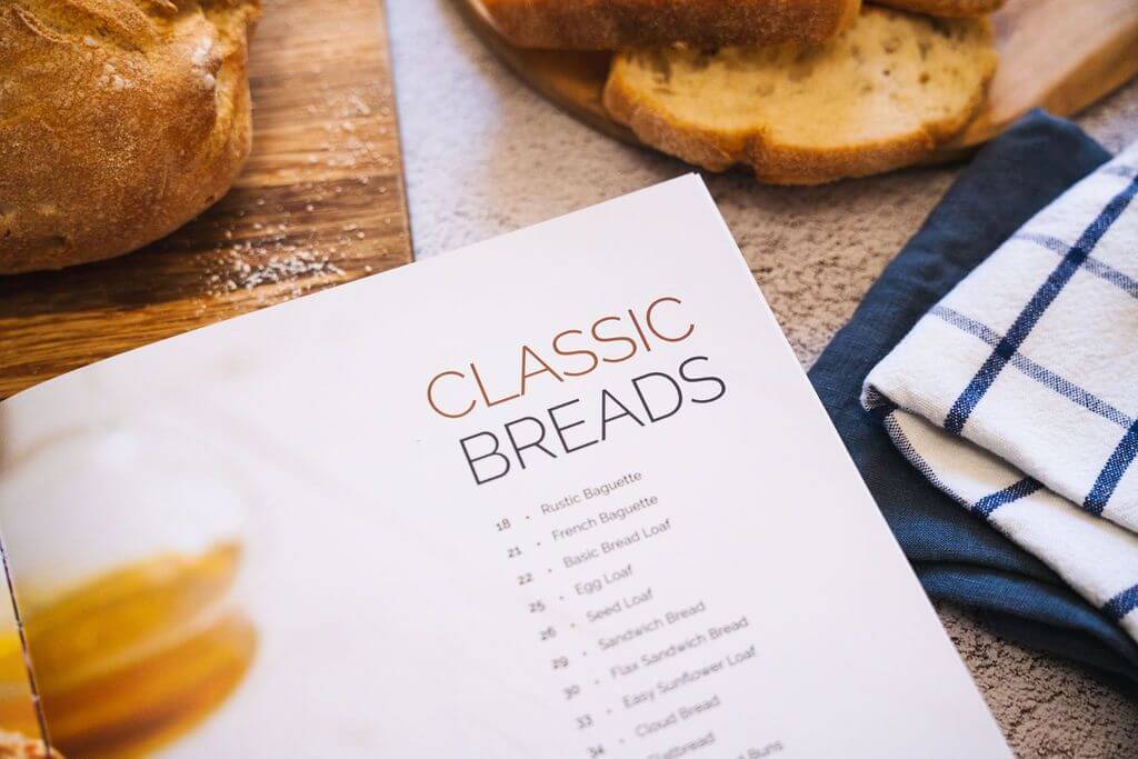 Wooden cutting board with bread ontop and the opened Keto Baker cookbook are placed on a marble surface.