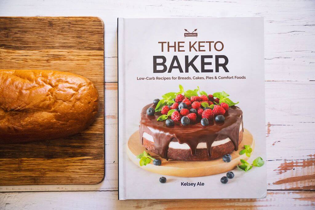 The Keto Baker book is placed on a marble surface beside a wooden board with bread.