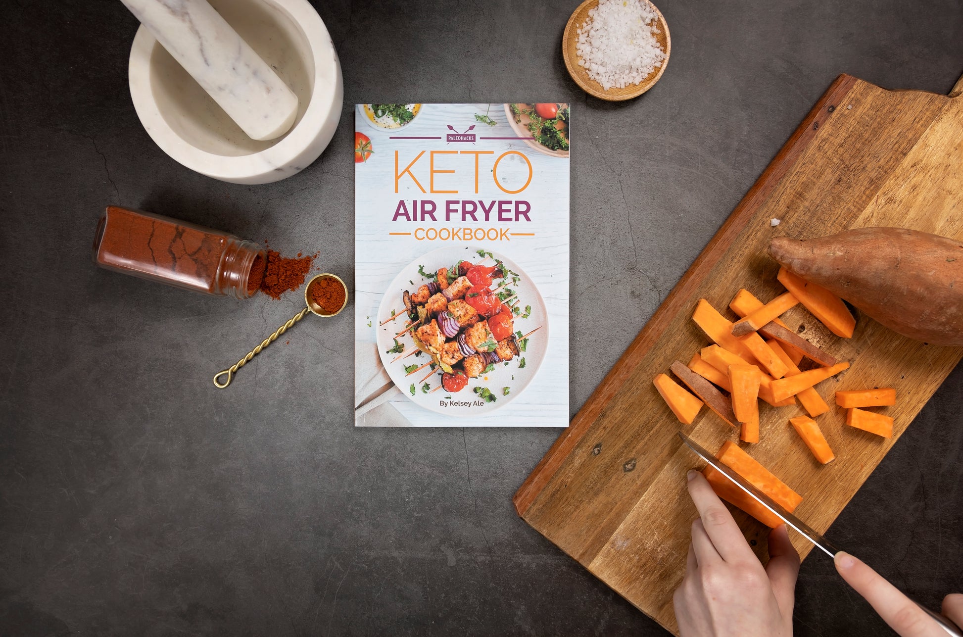 Keto air fryer cookbook surrounded by a wooden board with diced sweet potatoes, a bottle of spice, sea salt, and a kitchenware.