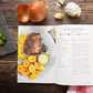 An opened Paleo Plenty Cookbook  laid on a wooden surface next to a chopping board with chopped peppers. Onions, garlic, and a kitchen cloth can also be seen.