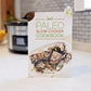 The Paleo Slow Cooker Cookbook stands on a marble kitchen counter. A pot and a bowl of fruit are seen in the background.
