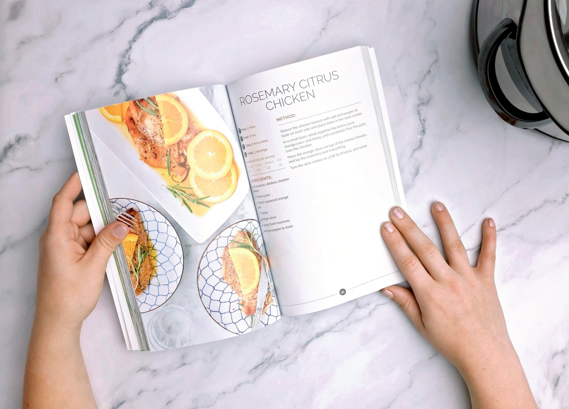 A woman holds an opened Paleo Slow Cooker Cookbook on a marble surface.