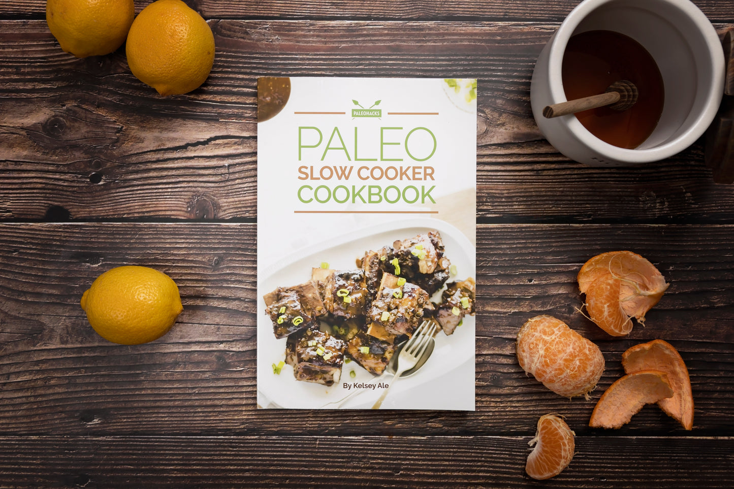 Paleo Slow Cooker Cookbook placed on a wooden surface with lemons and a jar of honey.
