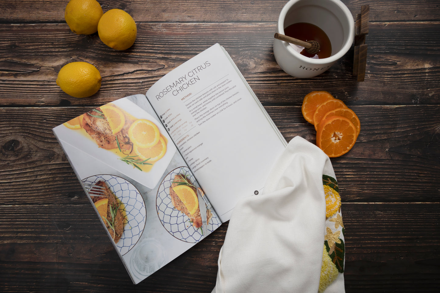 An opened Paleo Slow Cooker Cookbook on a wooden surface with lemon, a kitchen cloth and a jar of honey.