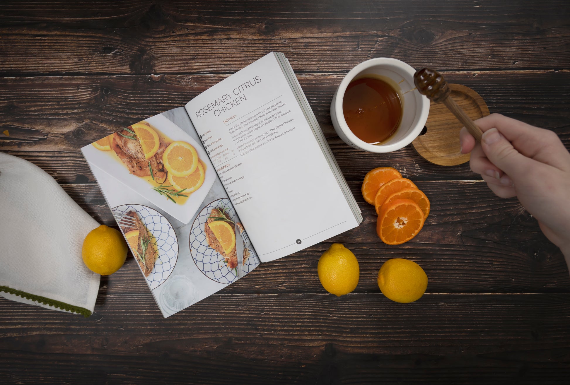 An opened Paleo Slow Cooker Cookbook laid on a wooden surface with citrus fruits and a jar of honey.
