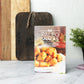 Paleo Snacks Cookbook stands on a marble kitchen counter with chopping boards and a flower pot.