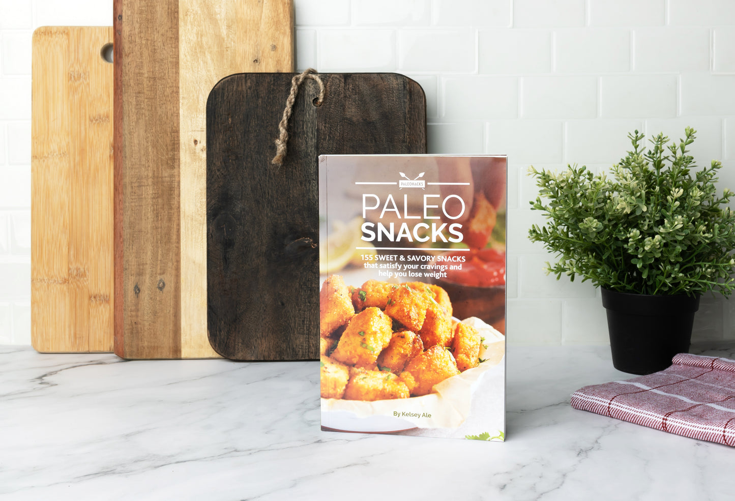 Paleo Snacks Cookbook stands on a marble kitchen counter with chopping boards and a flower pot.