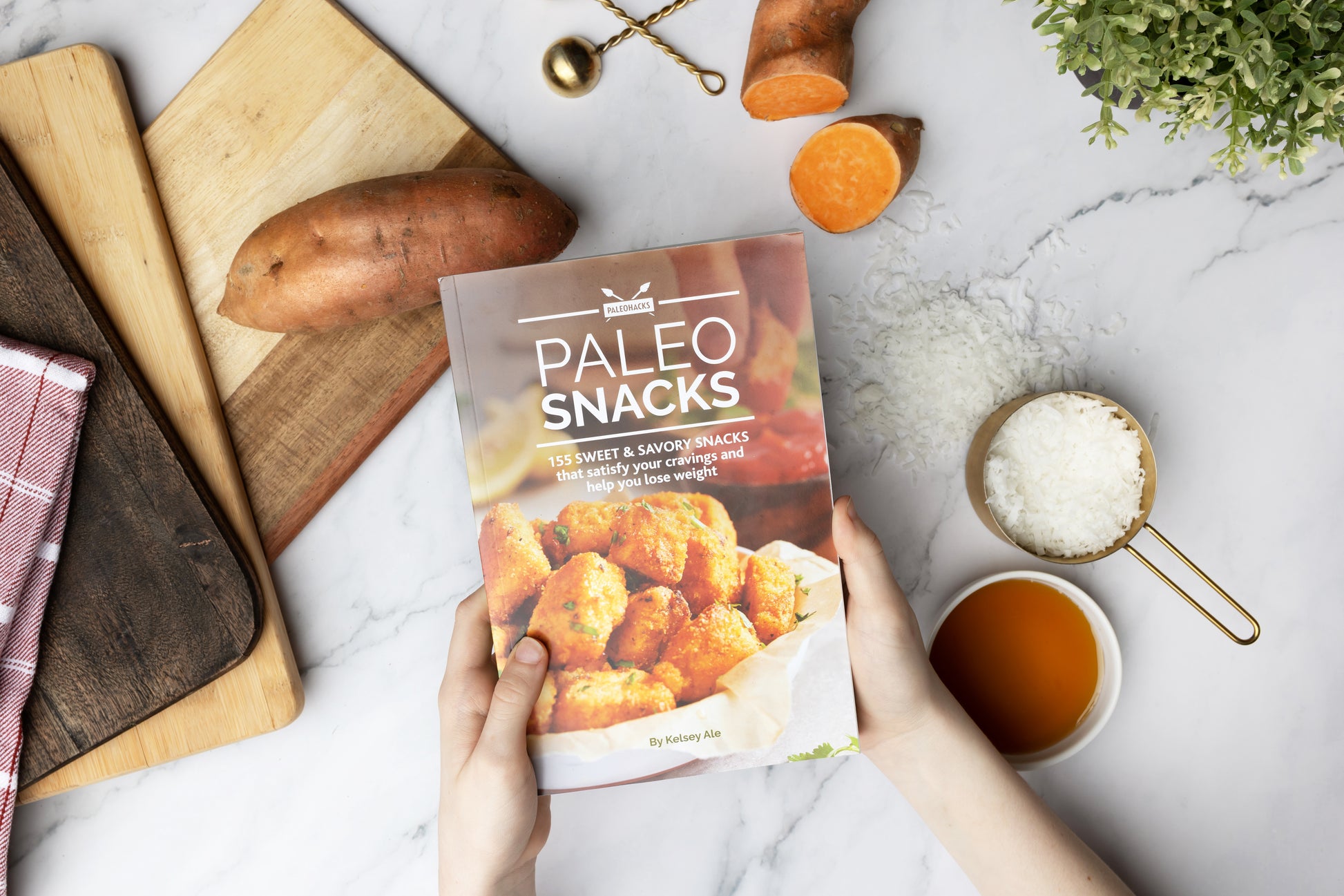 A woman holds the Paleo Snacks Cookbook above a marble surface with cutting boards, potatoes, vegetables, sea salt, and honey.