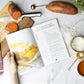 A woman holding the Paleo Snacks Cookbook, placed on a marble surface next to a cutting board, a knife, potatoes, shredded coconut, a kitchen cloth, and a bowl of honey.
