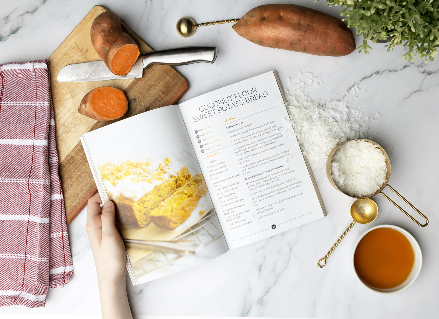 A woman holding the Paleo Snacks Cookbook, placed on a marble surface next to a cutting board, a knife, potatoes, shredded coconut, a kitchen cloth, and a bowl of honey.