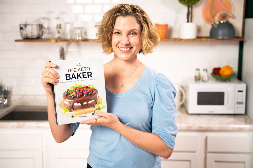 The cookbook author Kelsey Ale stands in the kitchen, holding the Keto Baker recipe book.