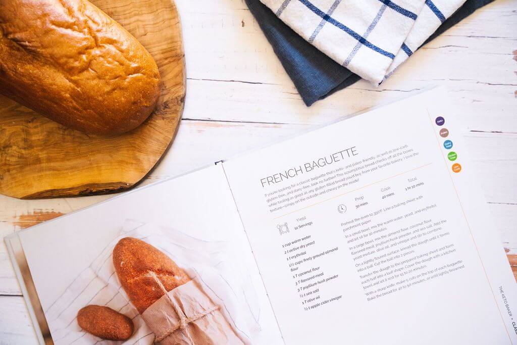 Opened Keto Baker cookbook placed on a marble surface next to a wooden board with bread and a kitchen cloth.