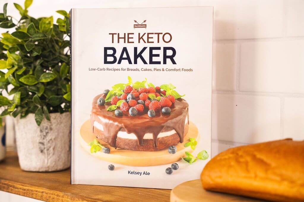 The Keto Baker cookbook leans against a white tile wall. A flower pot and bread can be seen on the wooden plain surface.